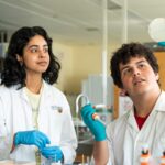 Two students, wearing lab coats, are working. One holds up a petri dish.