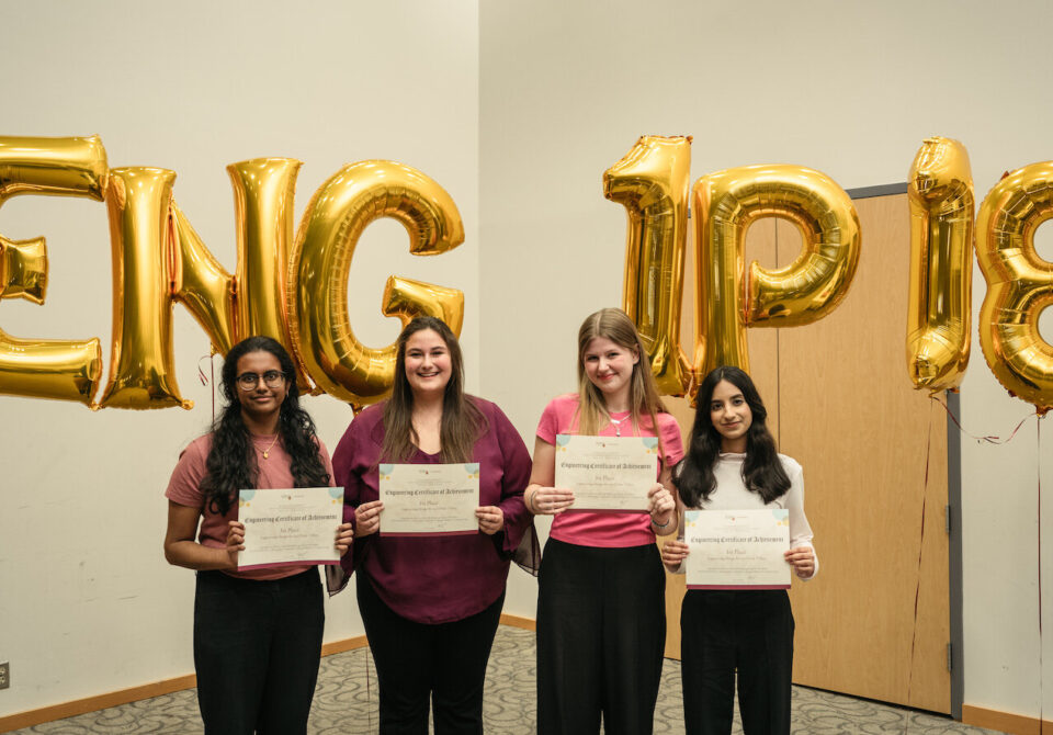 Four people standing in front of gold foil balloons holding up certificates.