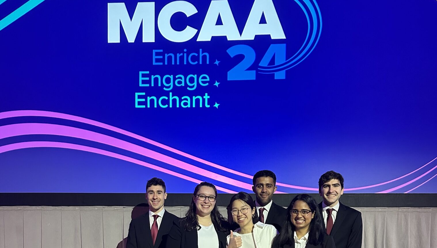 group of six students in business formal outfits posing in front of a powerpoint slide.