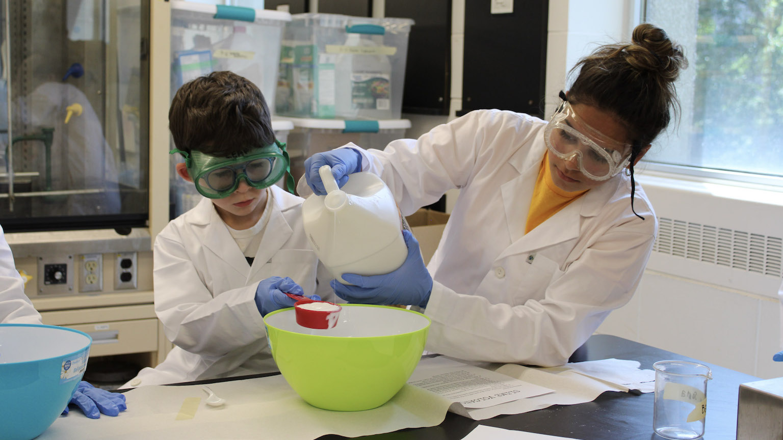 a young student and a camp instructor working on a science experiment in a lab. They are both wearing safety goggles and lab coats.