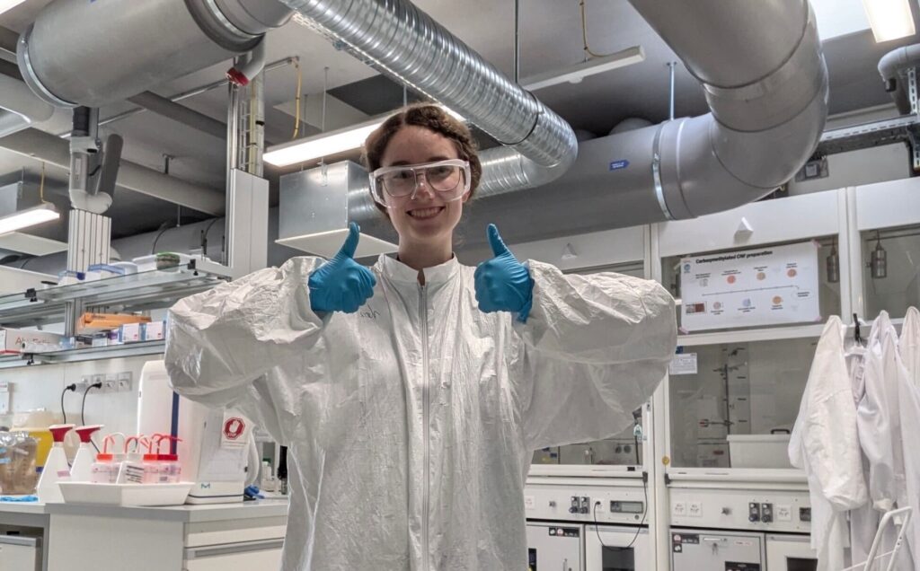 a woman in a lab wearing a clean suit, safety goggles and blue gloves.