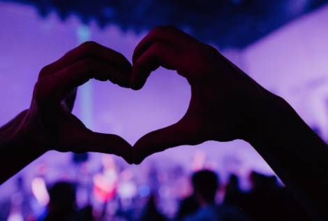 Heart hands in the foreground with concert goers and purple lights in the background