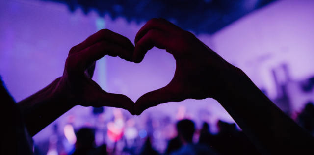 Heart hands in the foreground with concert goers and purple lights in the background