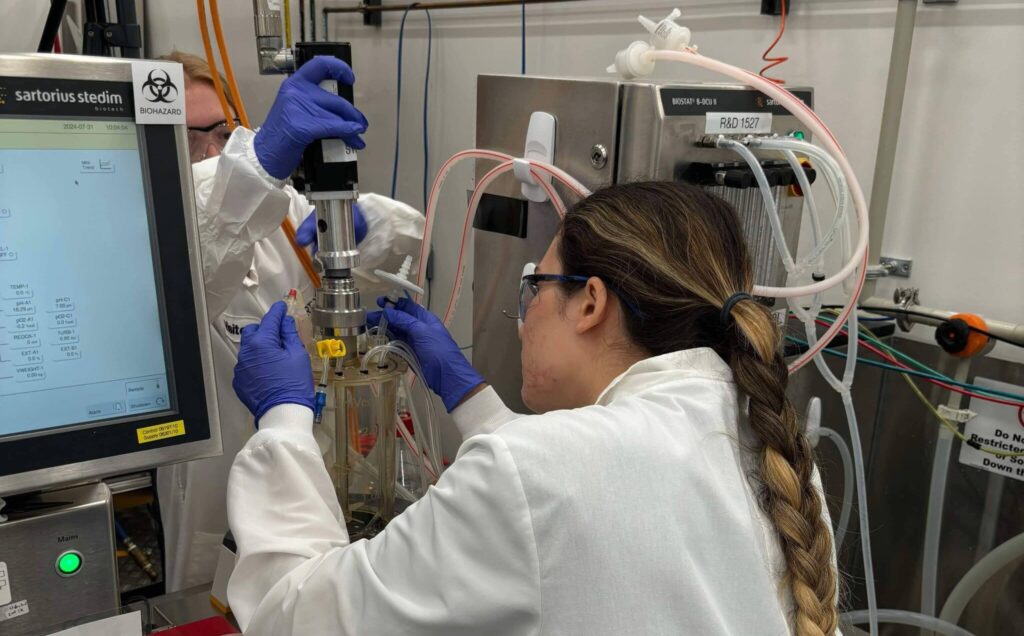 A woman with a braided ponytail and blue gloves is operating laboratory equipment alongside another individual. They are working with a bioreactor setup, carefully adjusting tubes connected to a stainless-steel apparatus. The setup includes a control screen displaying scientific data.