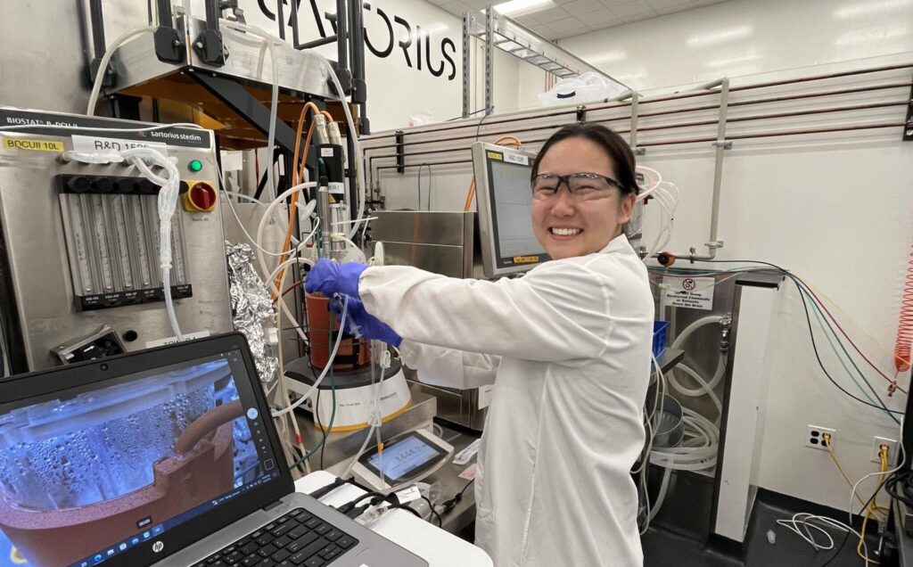 A woman in a lab coat and safety goggles is smiling while working in a laboratory setting. She is wearing blue gloves and adjusting equipment connected to a bioreactor system with tubes and sensors. A laptop in front of her shows a close-up of a bubbling liquid inside a flask.