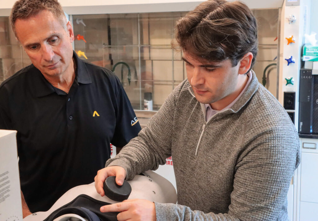 Carson Brewer simulates a hockey puck hitting a protective neck guard. Joe Camillo stands beside him.