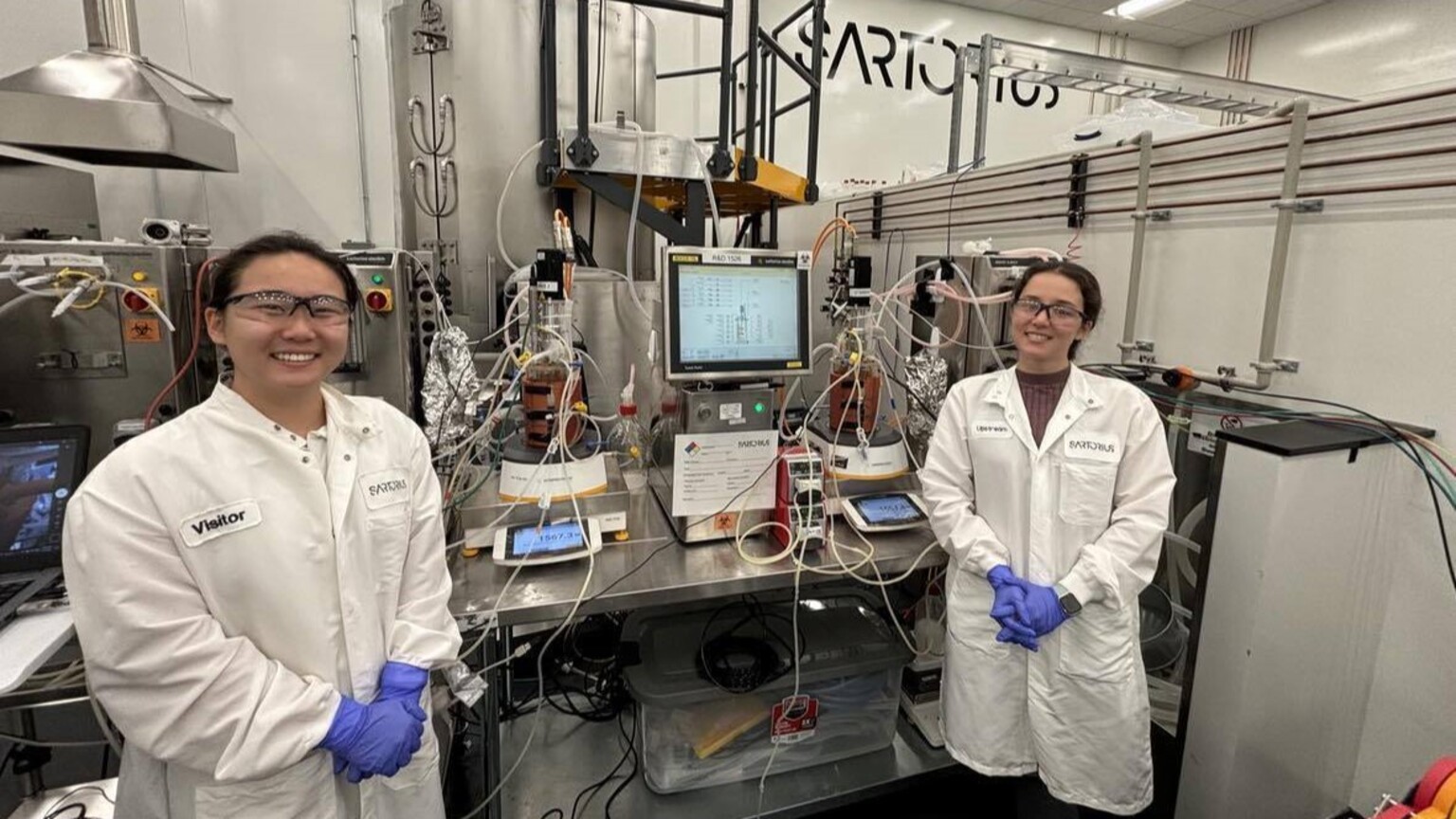 Two women in lab coats and safety goggles stand in front of a bioreactor system in a laboratory. They are surrounded by wires and tubing, smiling for the camera. The equipment in the background displays a control panel with monitoring data for their experiment.