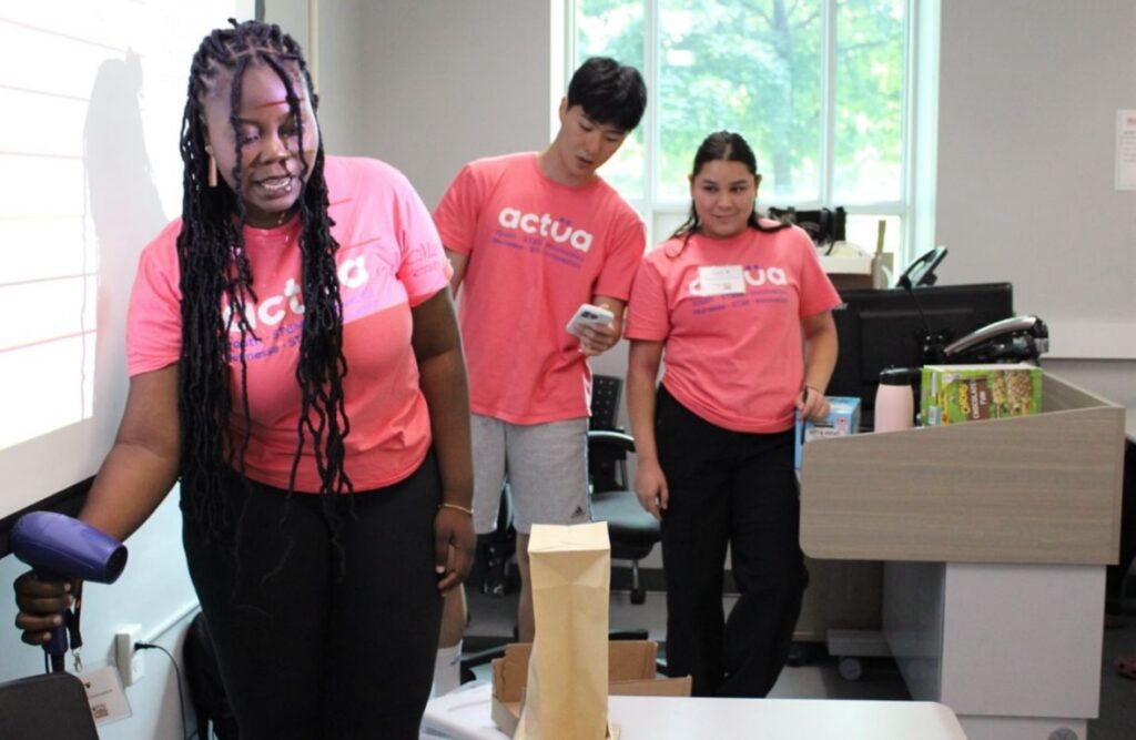 three activity facilitators wearing pink t-shirts standing at the front of a room while one of them is demonstrating a project by holding up a hair dryer to a brown paper lunch bag. 