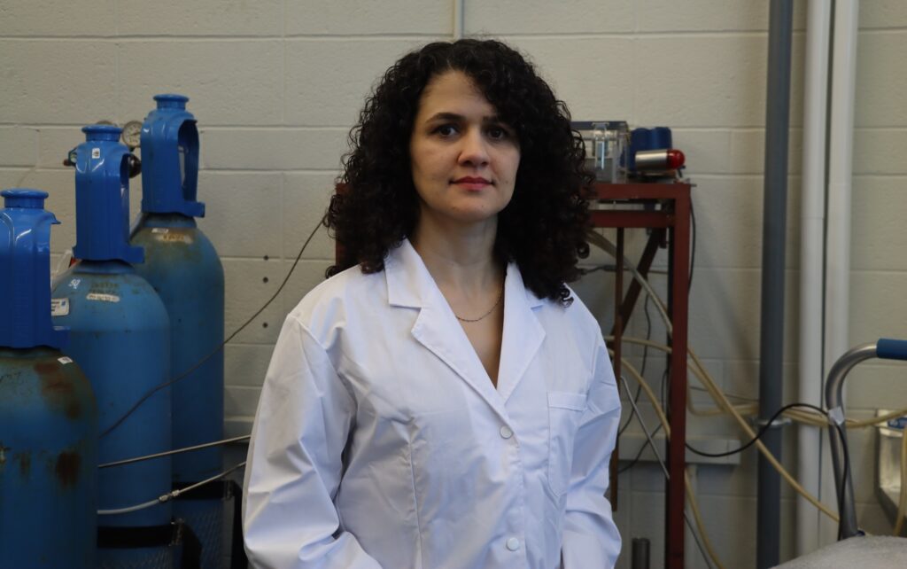 a woman in a white lab coat posing in front of blue hydrogen tanks.