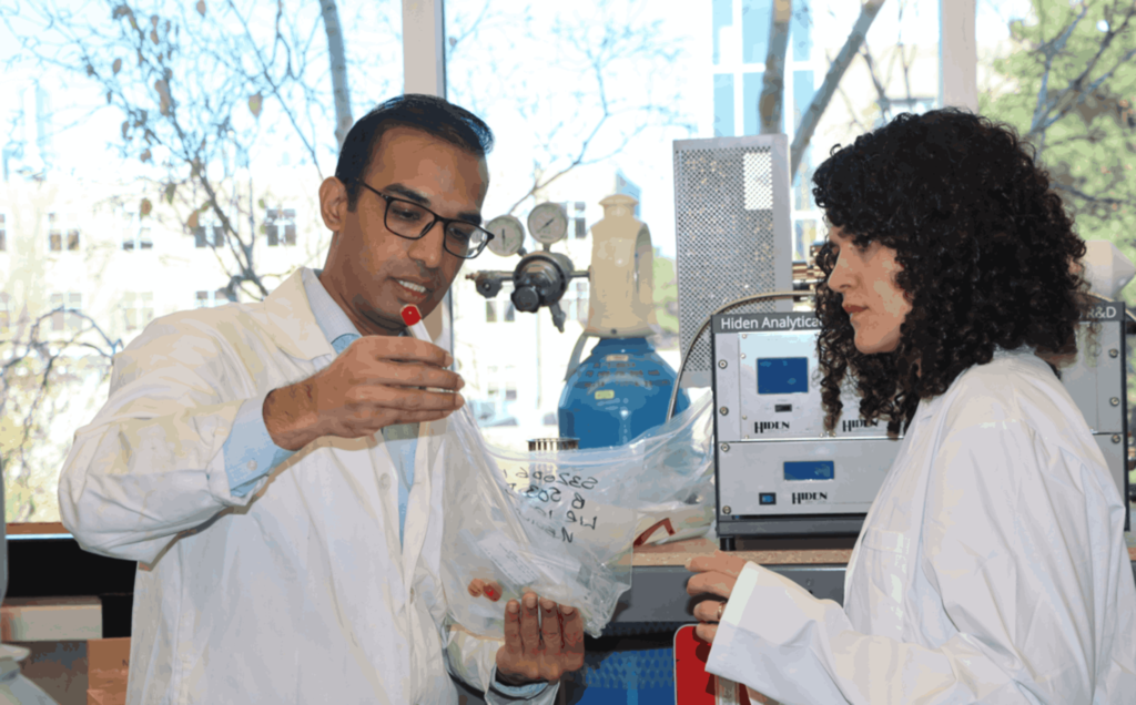 A man in a white lab coat, wearing glasses, holds a small red item while discussing it with a woman in a white lab coat with curly hair. They are in a laboratory with various scientific equipment in the background, including gas tanks and analytical devices.