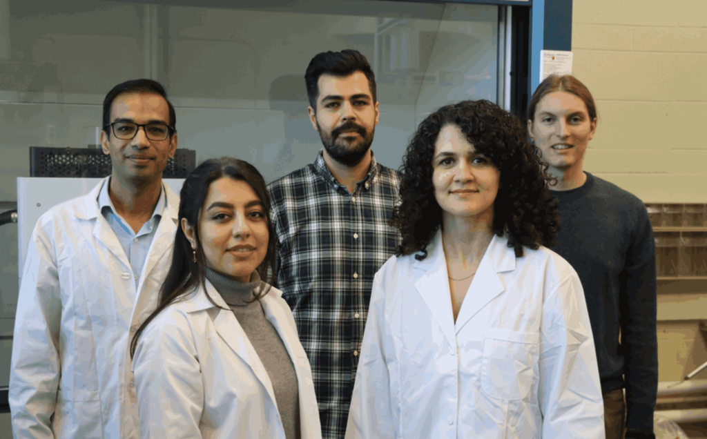 A group of five individuals standing in a lab environment. Two women in white lab coats are in the foreground, while three men, one in a checkered shirt and two in casual clothing, are in the background. They appear to be part of a scientific team.